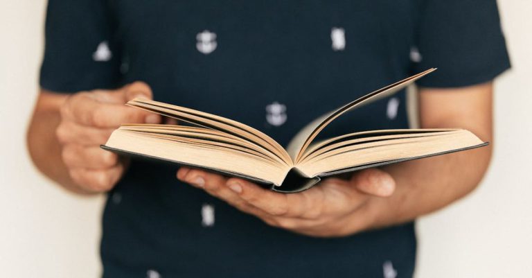 Literacy - Anonymous male in dark tee shirt reading interesting book while standing in front of light wall